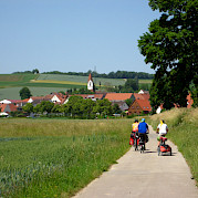 Tauber River Valley Romantic Road - Cyclists riding down a paved path through scenic countryside towards a village with red-roofed houses and a prominent church tower in Bavaria, Germany.