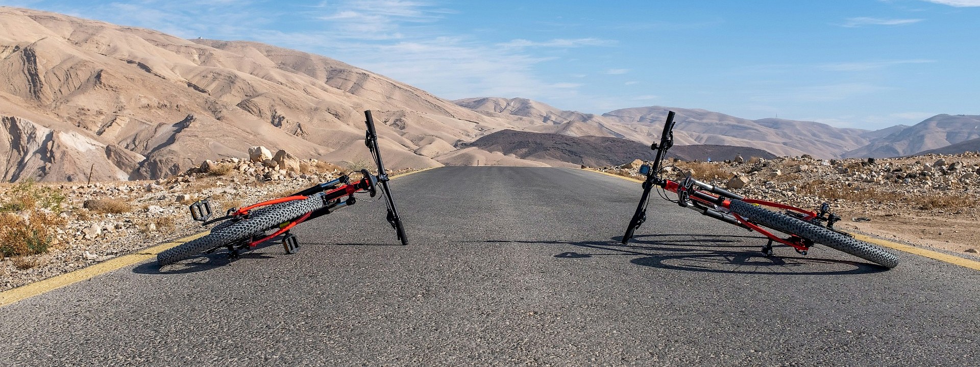 Two bicycles are lying on an open road in a barren, mountainous landscape under a clear blue sky.