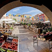 Hidden Treasures of the French Riviera - A vibrant market square in a French village, viewed through an archway. Stalls display fresh produce, and colorful flags adorn the open space, surrounded by tables with umbrellas and pastel-colored buildings.