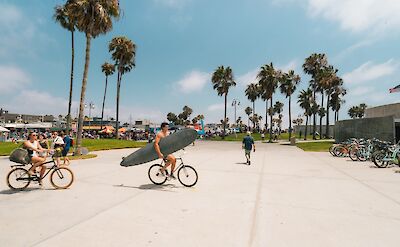 Biking while holding a surf board, Venice beach, California, USA. Unsplash: Matthew LeJune