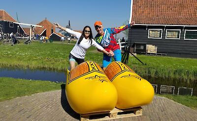 Two people stand inside giant yellow wooden clogs, with traditional Dutch houses in the background and a clear blue sky overhead.
