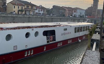 The boat "Ave Maria" travels along a canal at sunset, with a church and houses in the background.