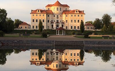 A large, elegant mansion with multiple windows and orange roofs, reflected in a calm pond in the foreground. The setting features a neatly landscaped garden.