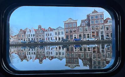 Row of historic European houses reflected in a canal, viewed through a window.