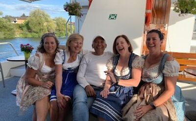 A group of five people sitting together, smiling on a boat deck. Four of them are wearing traditional dresses, and the setting suggests a casual, festive atmosphere.