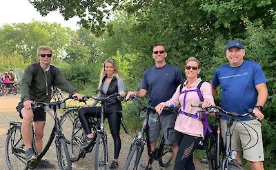 A group of five people standing with their bicycles on a paved path, surrounded by greenery. They are dressed in casual sportswear and appear ready for a cycling tour.