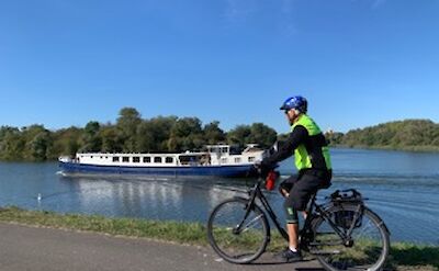 A person cycling along a path beside a river, while a long riverboat passes by on the water. The sky is clear and blue, creating a scenic, peaceful setting.