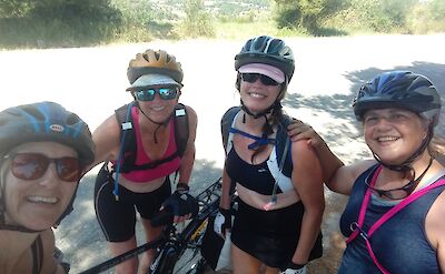 Four women wearing cycling gear and helmets, smiling for a photo while standing with a bicycle on a sunny road. The background shows greenery and a distant landscape.