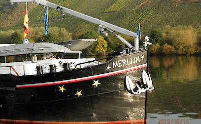 The bow of the boat "Merlijn" with star decorations, docked near a vineyard-covered hillside.