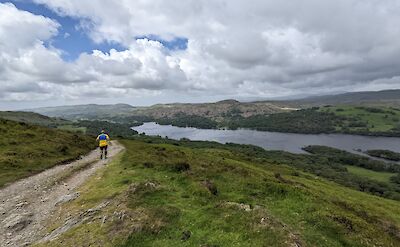A person walking along a grass and dirt path on a hill overlooking a large lake and distant hills under a cloudy sky.