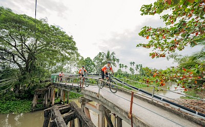 Cyclists cross a narrow rural bridge over a Mekong canal. toMBS
