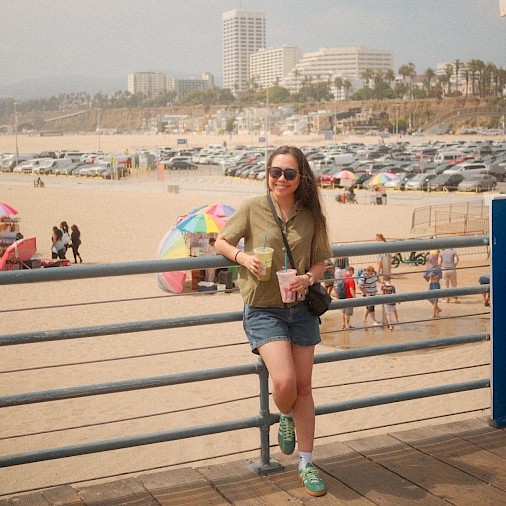 A person wearing sunglasses and holding two drinks stands on a promenade with a beach, colorful umbrellas, and city buildings in the background.