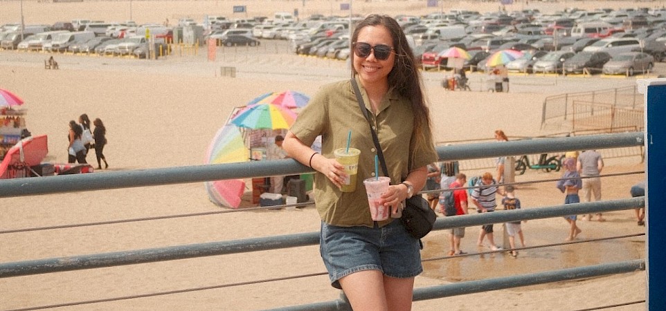 A person wearing sunglasses and holding two drinks stands on a promenade with a beach, colorful umbrellas, and city buildings in the background.